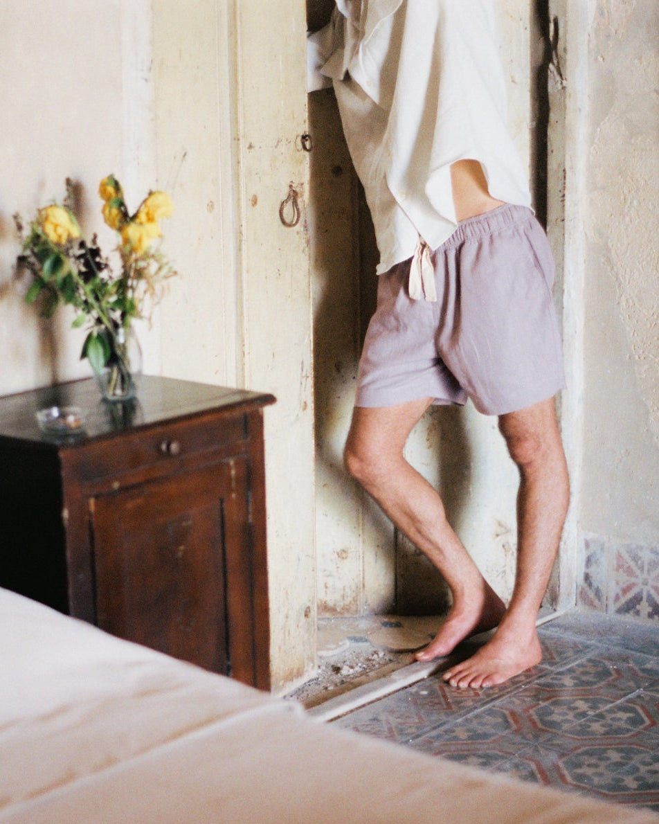 a man stadning inside a room catching a glance wearing a pair of linen shorts and white linen shirt, next to him a vase full of dead yellow roses and an ashtray with a smoking cigarrette.