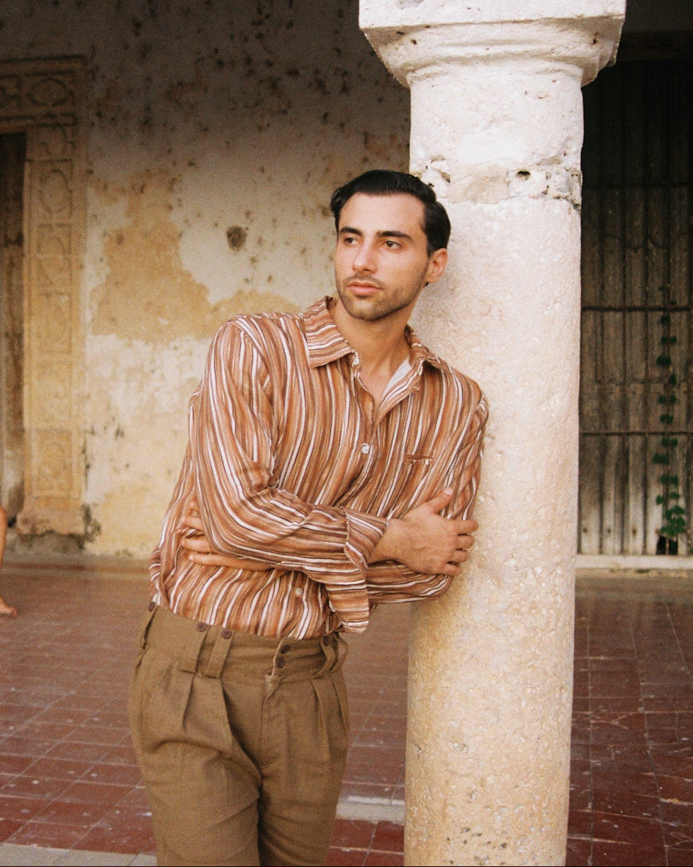 A man standing in front of an abandoned hacienda wearing a linen shirt and shorts, reclining next to a pilar with a serene face and beard, the outfit resembles a traveler's style. 