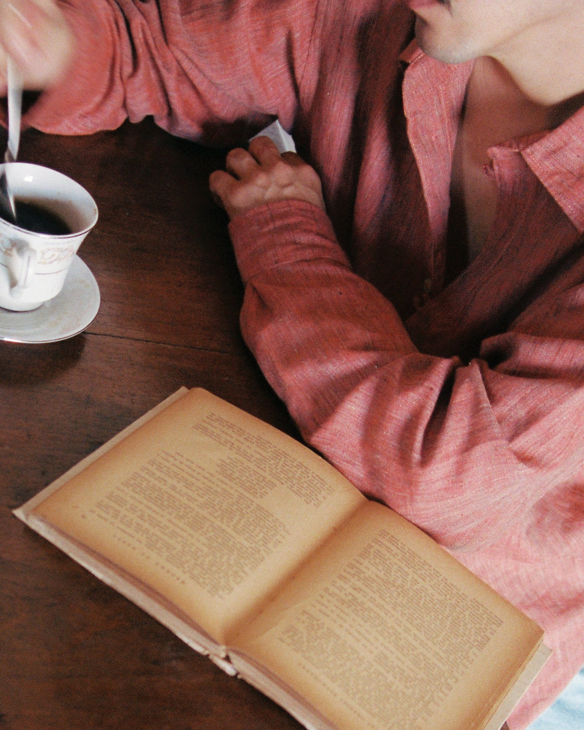 A man playing with a cup of coffee, a spoon rests inside of the black liquid, he's wearing a terracota linen shirt and a yellowed-page book lies resting on a table in front of him. 